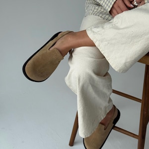 Könnte beinhalten: Beigefarbene Wildleder-Slipper mit schwarzer Sohle. Die Schuhe sind neutralfarben und haben ein schlichtes, klassisches Design. Die Schuhe werden an einer Person gezeigt, die auf einem Holzschemel sitzt.