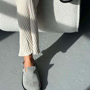 May include: A pair of gray suede clogs with a black sole. The clogs have a buckle closure and are worn with cream-colored ribbed pants. The image is taken in natural light, casting a shadow on the gray floor.