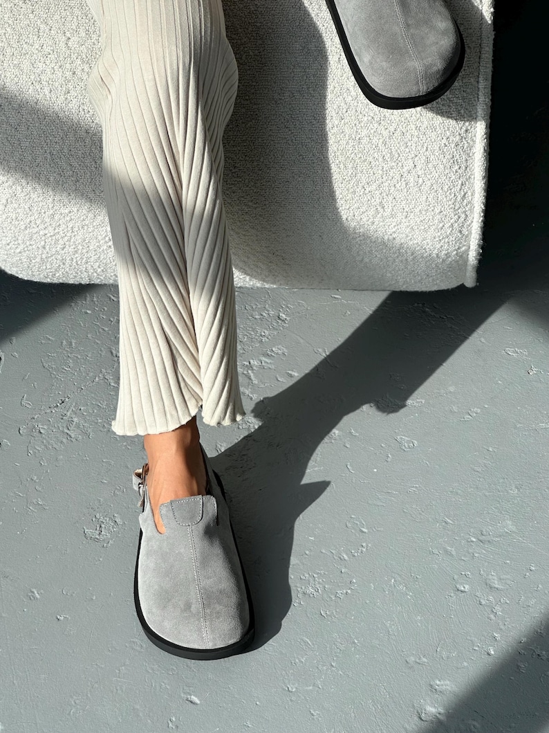 May include: A pair of gray suede clogs with a black sole. The clogs have a buckle closure and are worn with cream-colored ribbed pants. The image is taken in natural light, casting a shadow on the gray floor.
