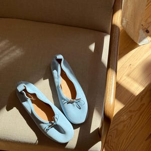 May include: A pair of light blue ballet pumps featuring a soft suede-like texture, a rounded toe, and a delicate bow detail at the front with metallic silver-toned aglets. The interior of the shoes is lined with a tan material and includes a signature.