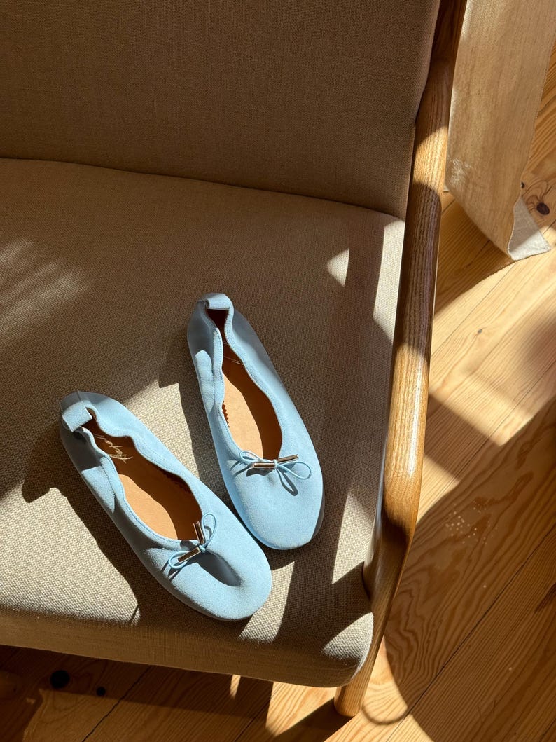 May include: A pair of light blue ballet pumps featuring a soft suede-like texture, a rounded toe, and a delicate bow detail at the front with metallic silver-toned aglets. The interior of the shoes is lined with a tan material and includes a signature.