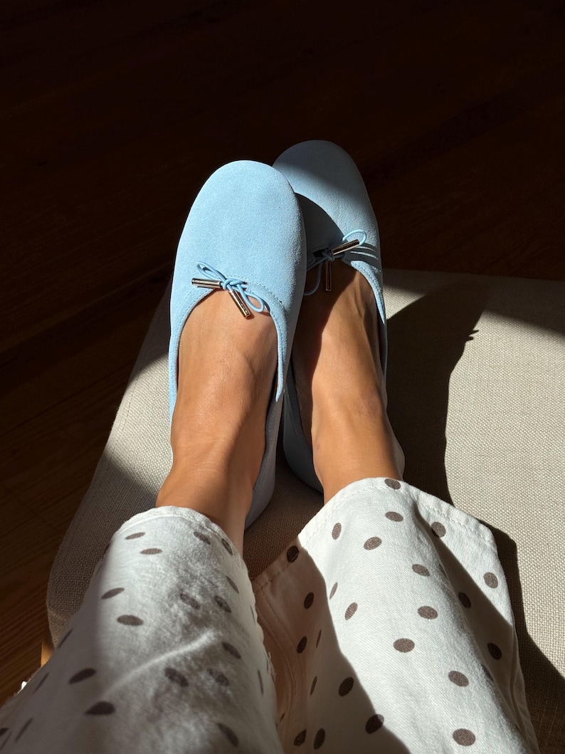 May include: A pair of light blue suede ballet flats featuring a rounded toe and a small decorative bow at the front with metallic gold-toned aglets. The shoes are worn with white trousers that have a brown polka dot pattern throughout the fabric.