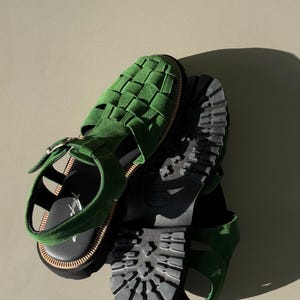 Puede incluir: Sandalias de ante verde con dise&ntilde;o superior tejido. Las sandalias tienen una suela negra con dibujo de tacos y cierre de hebilla. Las sandalias se muestran desde arriba.