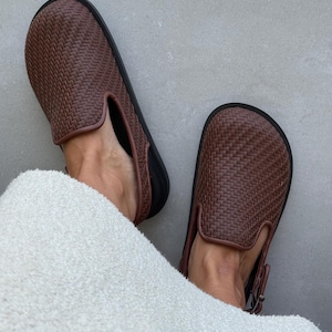 May include: Brown woven leather slip-on shoes with black soles. The shoes have a classic design with a textured pattern. The image shows the shoes on a light gray surface.