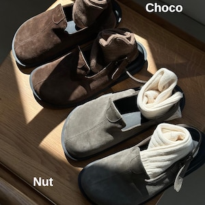 May include: Close-up of two pairs of suede slip-on shoes in brown and gray, each with a pair of rolled socks inside. The shoes are on a wooden surface. The text "Choco" and "Nut" are visible.