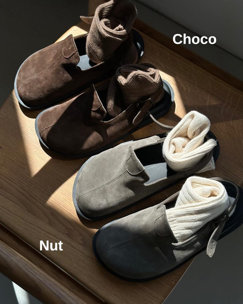 May include: Close-up of two pairs of suede slip-on shoes in brown and gray, each with a pair of rolled socks inside. The shoes are on a wooden surface. The text "Choco" and "Nut" are visible.
