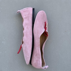 May include: A pair of light pink suede ballet flats featuring a scalloped topline, a thin red elastic cord detail across the vamp, and a small gold-toned metal aglet. The shoes have a flat black sole and a small pull tab at the heel counter.