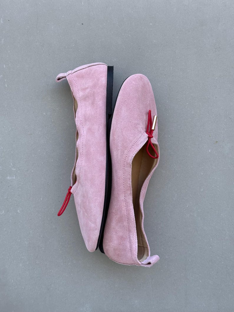 May include: A pair of light pink suede ballet flats featuring a scalloped topline, a thin red elastic cord detail across the vamp, and a small gold-toned metal aglet. The shoes have a flat black sole and a small pull tab at the heel counter.