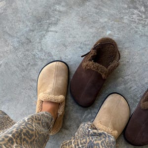 Könnte beinhalten: Zwei Paar Wildleder-Hausschuhe. Ein Paar ist beige mit schwarzer Sohle und braunem Shearling-Futter. Das andere Paar ist braun mit Shearling-Futter und einem Riemen. Auf einer grauen Betonoberfläche.