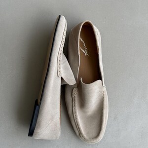 Puede incluir: Un par de mocasines de ante beige claro. Los zapatos tienen una puntera redondeada, una pala cosida y una suela negra. El interior del zapato tiene un forro de cuero marr&oacute;n con un logotipo impreso.