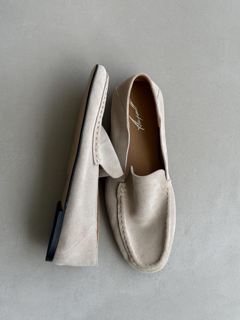 Puede incluir: Un par de mocasines de ante beige claro. Los zapatos tienen una puntera redondeada, una pala cosida y una suela negra. El interior del zapato tiene un forro de cuero marr&oacute;n con un logotipo impreso.
