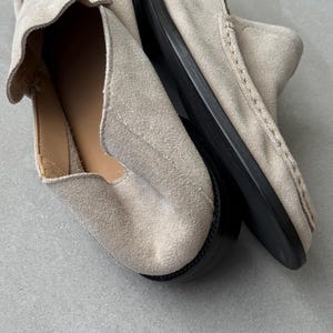 May include: A pair of light beige suede loafers with a rounded toe and a black sole. The shoes have a slip-on design and visible stitching details. The interior lining is a light tan color.