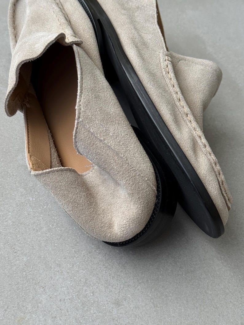 May include: A pair of light beige suede loafers with a rounded toe and a black sole. The shoes have a slip-on design and visible stitching details. The interior lining is a light tan color.