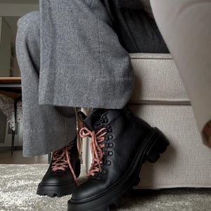May include: Black leather boots with thick, lug soles and pink laces. The boots have a lace-up design with metal eyelets. The boots are worn with gray pants. The boots are on a gray rug.