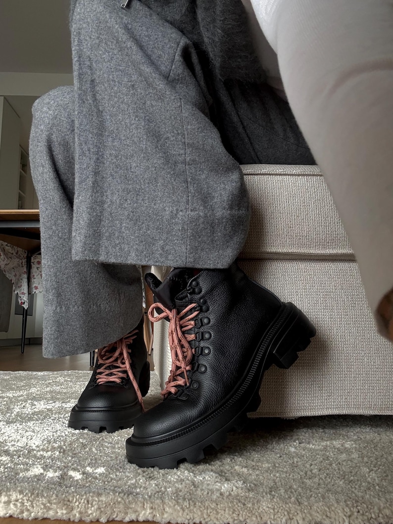 May include: Black leather boots with thick, lug soles and pink laces. The boots have a lace-up design with metal eyelets. The boots are worn with gray pants. The boots are on a gray rug.