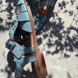 K&ouml;nnte beinhalten: Ein Paar hellblaue Ledersandalen mit gewebtem Design. Die Sandalen haben verstellbare Schnallen und eine beige Sohle. Die Sandalen sind etwa 2.5 cm hoch.