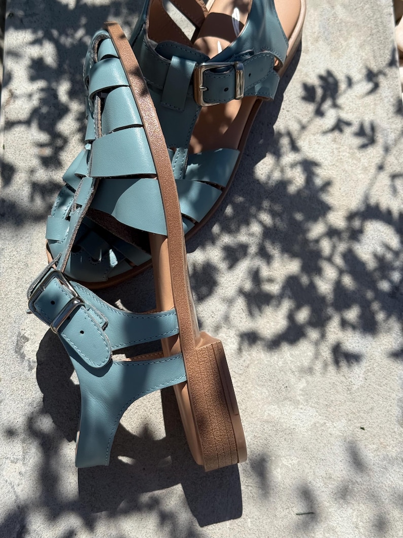 K&ouml;nnte beinhalten: Ein Paar hellblaue Ledersandalen mit gewebtem Design. Die Sandalen haben verstellbare Schnallen und eine beige Sohle. Die Sandalen sind etwa 2.5 cm hoch.