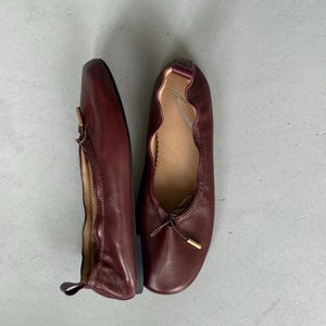 May include: A pair of dark burgundy leather ballet flats featuring a rounded toe, a gathered elastic topline, and a thin decorative bow at the front with gold-toned metal aglets. The interior sole is tan and displays a cursive brand logo.