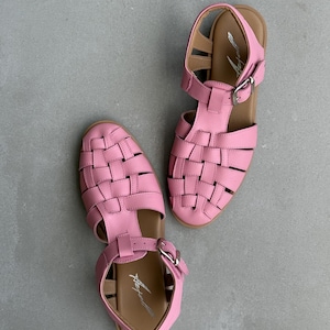 K&ouml;nnte beinhalten: Ein Paar rosa Ledersandalen mit geflochtenem Design an der Spitze und Schnallenverschluss. Die Sandalen haben eine braune Sohle und sind vor grauem Hintergrund platziert. Sommerschuhe.