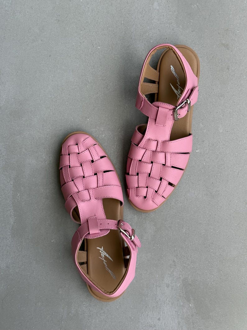 K&ouml;nnte beinhalten: Ein Paar rosa Ledersandalen mit geflochtenem Design an der Spitze und Schnallenverschluss. Die Sandalen haben eine braune Sohle und sind vor grauem Hintergrund platziert. Sommerschuhe.