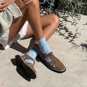 Puede incluir: Sandalias de cuero marr&oacute;n con correas tejidas y suela negra. Las sandalias se usan con calcetines azul claro. La imagen muestra las sandalias sobre una superficie de hormig&oacute;n.