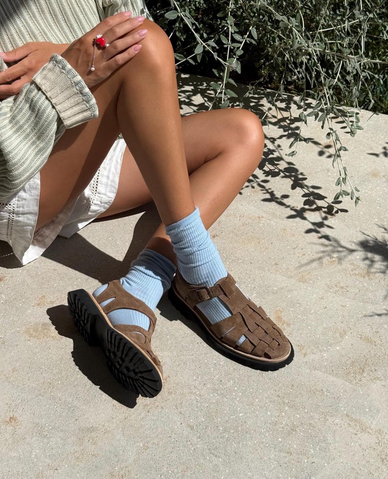 Puede incluir: Sandalias de cuero marr&oacute;n con correas tejidas y suela negra. Las sandalias se usan con calcetines azul claro. La imagen muestra las sandalias sobre una superficie de hormig&oacute;n.