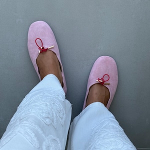 May include: A pair of light pink suede ballet flats featuring a thin red drawstring cord tied in a small bow at the centre of the vamp, accented with gold-toned metal aglets, paired with white textured trousers with a raised floral pattern.