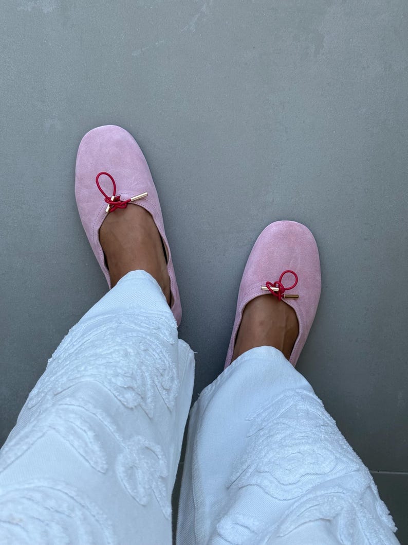 May include: A pair of light pink suede ballet flats featuring a thin red drawstring cord tied in a small bow at the centre of the vamp, accented with gold-toned metal aglets, paired with white textured trousers with a raised floral pattern.