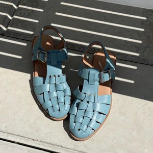 K&ouml;nnte beinhalten: Ein Paar hellblaue Ledersandalen mit einem gewebten Design. Die Sandalen haben verstellbare Kn&ouml;chelriemen mit silbernen Schnallen und braunen Sohlen. Sie liegen auf einer Betonoberfl&auml;che.