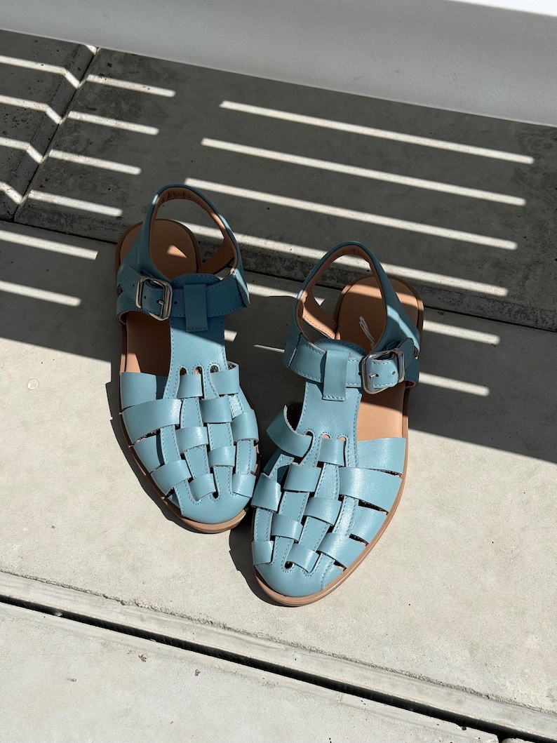 K&ouml;nnte beinhalten: Ein Paar hellblaue Ledersandalen mit einem gewebten Design. Die Sandalen haben verstellbare Kn&ouml;chelriemen mit silbernen Schnallen und braunen Sohlen. Sie liegen auf einer Betonoberfl&auml;che.