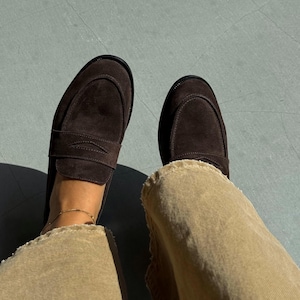 May include: A pair of dark brown suede loafers with a classic penny strap design. The shoes are worn with tan corduroy pants. The image is taken from a top-down perspective, showcasing the footwear and part of the outfit.