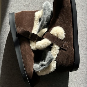 Può includere: Stivaletti alla caviglia in camoscio marrone con suola nera e chiusura con fibbia. Gli stivali hanno una soffice fodera color crema e un accento grigio. Gli stivali sono progettati per il comfort e il calore.
