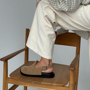 May include: A tan suede clog with a black sole and a buckle closure. The shoe is on a wooden chair. The clog has a rounded toe and a strap that goes around the ankle.