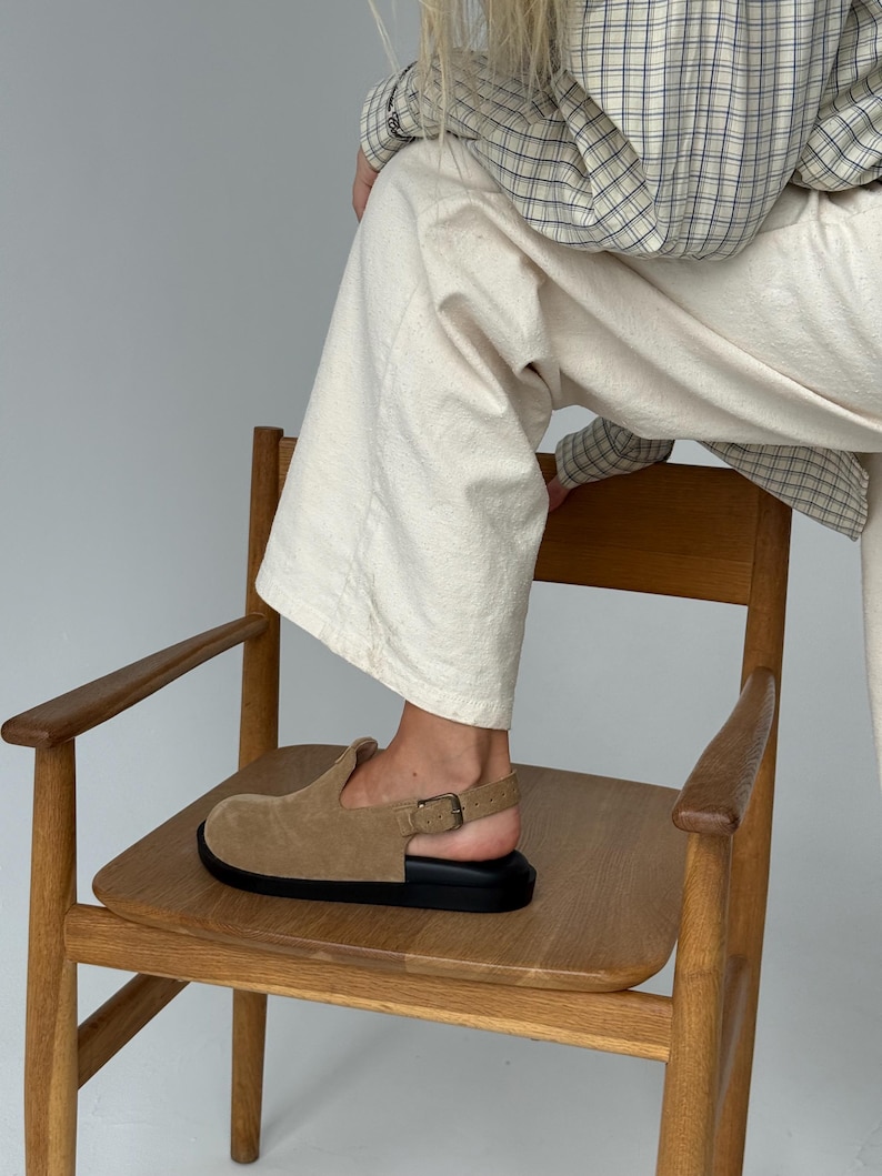 May include: A tan suede clog with a black sole and a buckle closure. The shoe is on a wooden chair. The clog has a rounded toe and a strap that goes around the ankle.