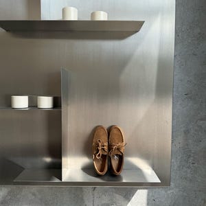 Op de afbeelding: Een paar bruine su&egrave;de bootschoenen met veters, uitgestald op een metalen plank. De schoenen zijn lichtbruin van kleur en hebben een klassiek ontwerp. De achtergrond toont een metalen muur en witte cilindrische objecten.
