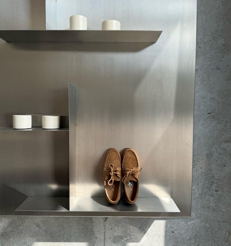 Op de afbeelding: Een paar bruine su&egrave;de bootschoenen met veters, uitgestald op een metalen plank. De schoenen zijn lichtbruin van kleur en hebben een klassiek ontwerp. De achtergrond toont een metalen muur en witte cilindrische objecten.