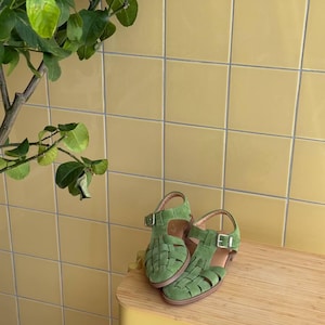 K&ouml;nnte beinhalten: Ein Paar gr&uuml;ne Wildleder-Sandalen mit geflochtenen Riemen und Schnallenverschl&uuml;ssen. Die Sandalen werden auf einer hellen Holzoberfl&auml;che vor einer gelben Fliesenwand pr&auml;sentiert. Die Sandalen sind leuchtend gr&uuml;n.