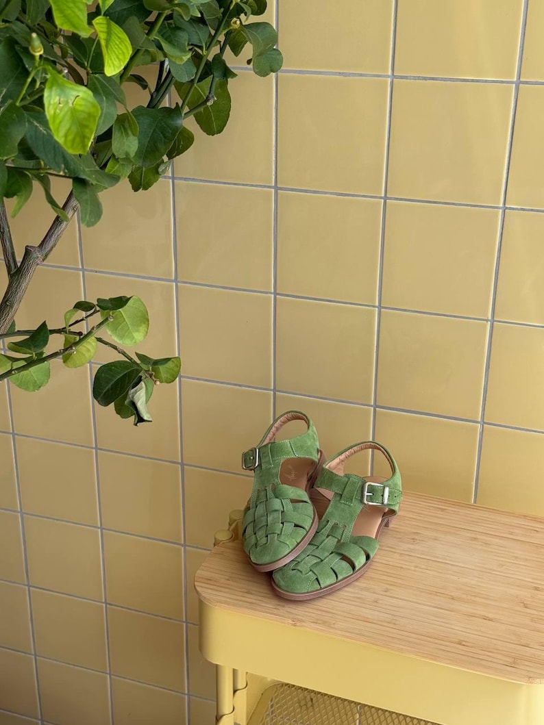 K&ouml;nnte beinhalten: Ein Paar gr&uuml;ne Wildleder-Sandalen mit geflochtenen Riemen und Schnallenverschl&uuml;ssen. Die Sandalen werden auf einer hellen Holzoberfl&auml;che vor einer gelben Fliesenwand pr&auml;sentiert. Die Sandalen sind leuchtend gr&uuml;n.