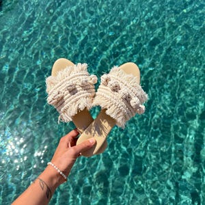 May include: A pair of beige macrame sandals with a cream-colored woven design. The sandals have a flat sole and are held over turquoise water. The sandals are a summer fashion item.