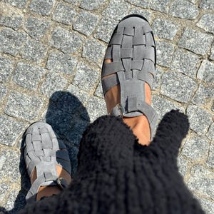 Puede incluir: Sandalias grises de ante con dise&ntilde;o tejido. Las sandalias tienen una tira en T y cierre de hebilla. La imagen est&aacute; tomada desde arriba, mostrando las sandalias sobre una superficie adoquinada.
