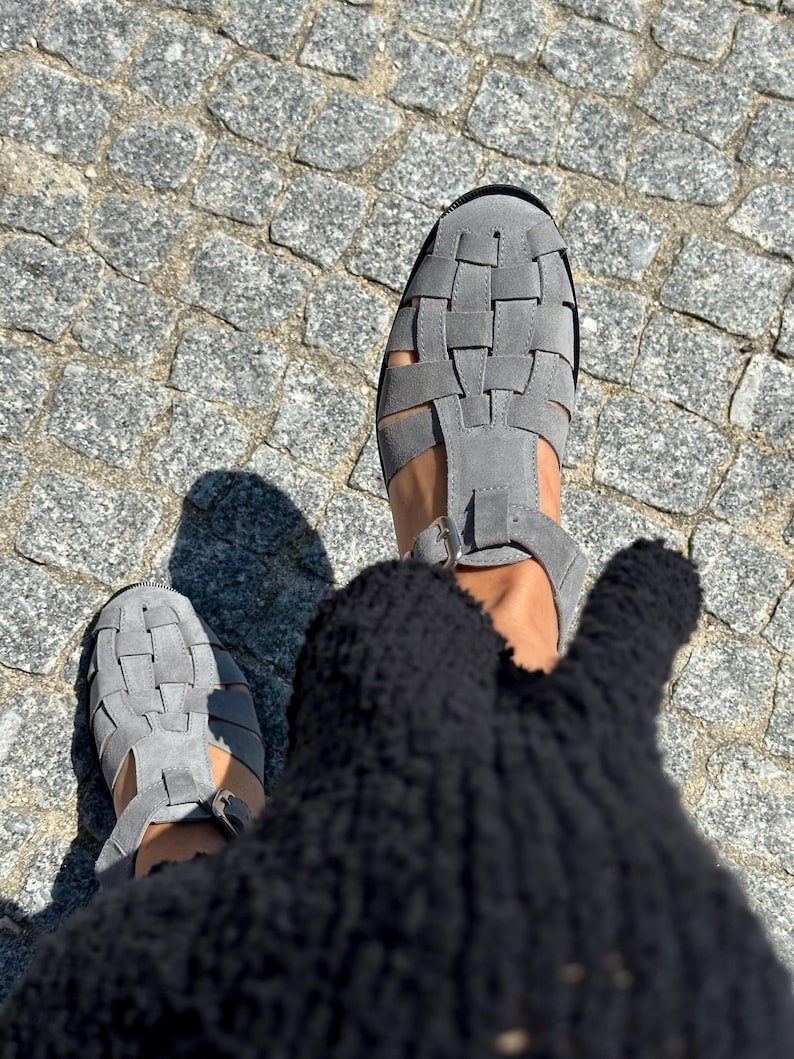 Puede incluir: Sandalias grises de ante con dise&ntilde;o tejido. Las sandalias tienen una tira en T y cierre de hebilla. La imagen est&aacute; tomada desde arriba, mostrando las sandalias sobre una superficie adoquinada.