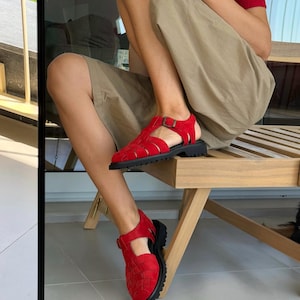Puede incluir: Sandalias rojas tejidas con suela negra y cierre de hebilla. Las sandalias tienen una suela gruesa y un dise&ntilde;o tejido en la parte superior. Las sandalias se usan en los pies de una persona sentada en una silla de madera.