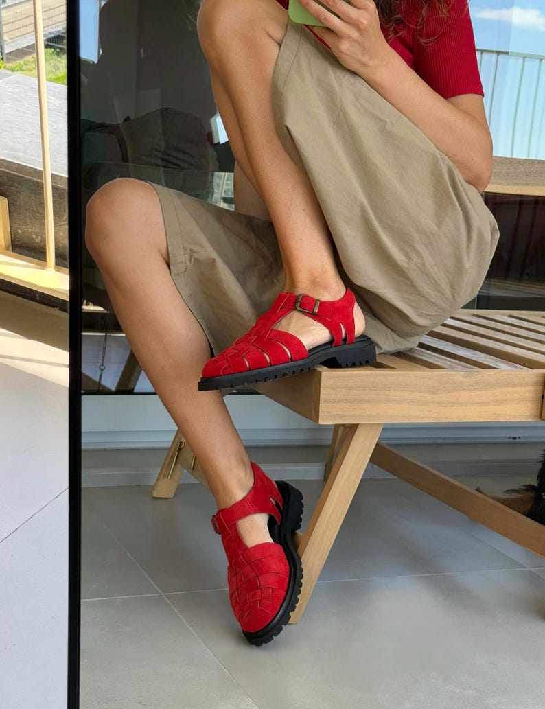 Puede incluir: Sandalias rojas tejidas con suela negra y cierre de hebilla. Las sandalias tienen una suela gruesa y un dise&ntilde;o tejido en la parte superior. Las sandalias se usan en los pies de una persona sentada en una silla de madera.