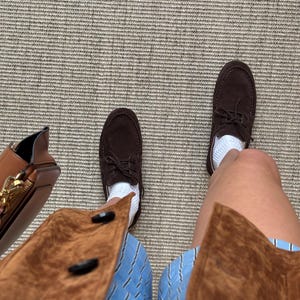 Op de afbeelding: Een bovenaanzicht toont bruine su&egrave;de bootschoenen met witte sokken, gecombineerd met een blauw-wit gestreepte short en een bruine su&egrave;de jas. Een bruine leren tas met een gouden rits is links zichtbaar.