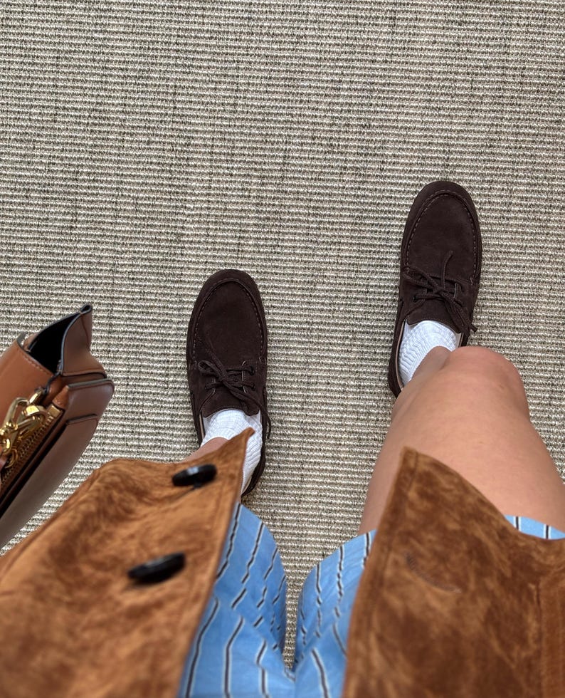 Op de afbeelding: Een bovenaanzicht toont bruine su&egrave;de bootschoenen met witte sokken, gecombineerd met een blauw-wit gestreepte short en een bruine su&egrave;de jas. Een bruine leren tas met een gouden rits is links zichtbaar.