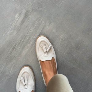May include: A pair of light beige suede loafers with tassel detailing. The shoes have a rounded toe and a low heel. The loafers are a neutral colour and appear to be unworn. The shoes are a casual style.