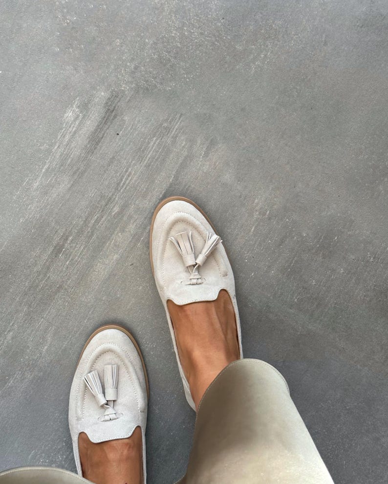 May include: A pair of light beige suede loafers with tassel detailing. The shoes have a rounded toe and a low heel. The loafers are a neutral colour and appear to be unworn. The shoes are a casual style.