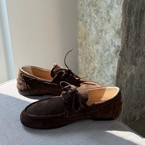Op de afbeelding: Een paar bruine su&egrave;de loafers met leren voering. De schoenen hebben een ronde neus, decoratieve stiksels en een geknoopt veterdetail. De loafers worden gepresenteerd op een grijze ondergrond.