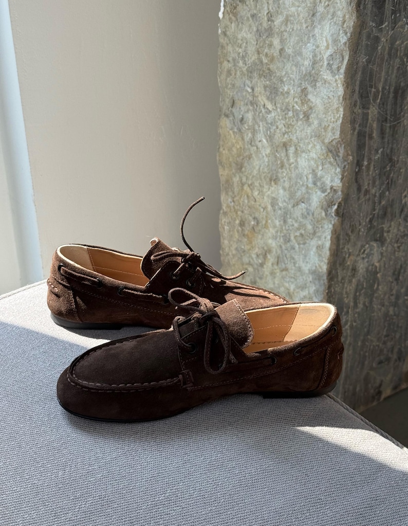 Op de afbeelding: Een paar bruine su&egrave;de loafers met leren voering. De schoenen hebben een ronde neus, decoratieve stiksels en een geknoopt veterdetail. De loafers worden gepresenteerd op een grijze ondergrond.