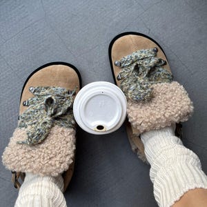 Könnte beinhalten: Hellbraune Wildleder-Clogs mit Kunstfellbesatz und Strickdetails. Die Clogs werden mit cremefarbenen Beinwärmern getragen. Ein weißer Kaffeetassendeckel liegt zwischen den Schuhen.